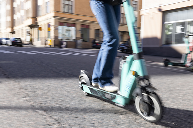 Ihminen ajaa vuokraamon turkoosilla sähköpotkulaudalla kaupungin katua. Kuvassa näkyy kuljettajasta vain farkut ja tennarit.