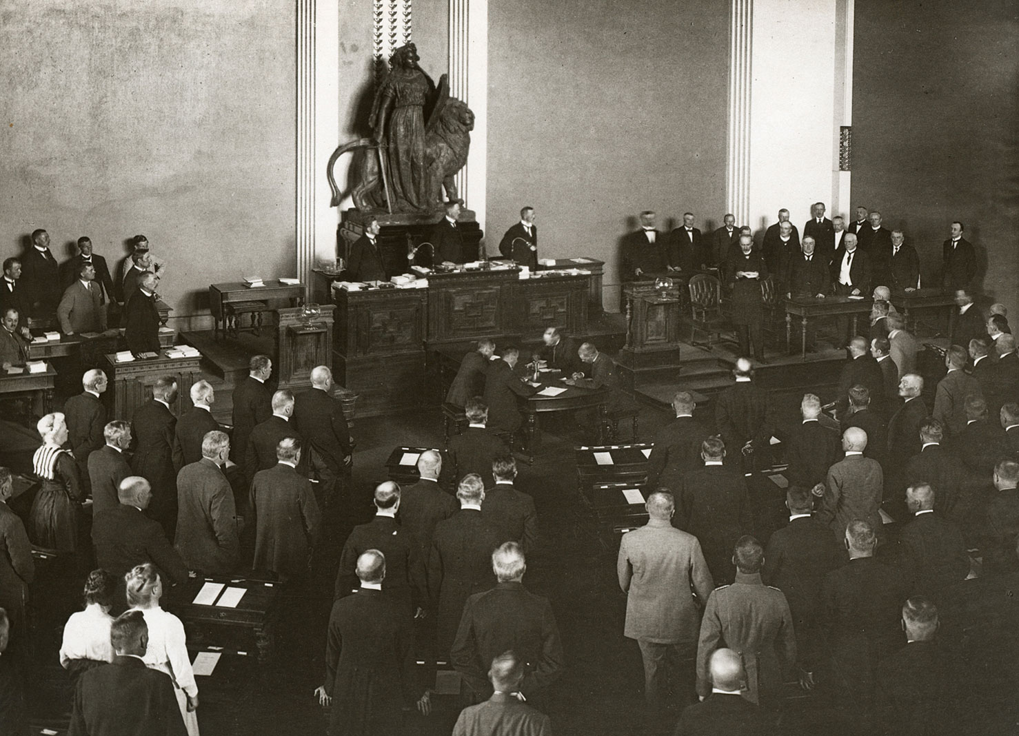 Valokuvassa tasavallan presidentti Kaarlo Juho Ståhlberg lausumassa juhlallinen vakuutustaan Heimolan talossa vuonna 1919. Osallistujat seisovat tilaisuutta kuuntelemassa. Etualalla salissa Walter Runebergin Lex-veistoksen kipsikopio, jossa on miekkaa ja kilpeä pitelevä oikeuden jumalatar, jonka takana on leijona.