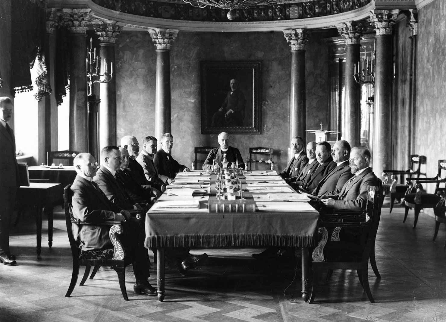 Valokuvassa A.K. Cajanderin virkamieshallitus presidentin esittelyssä vuonna 1922. Pöydän päässä presidentti K.J. Ståhlberg. Huoneen takaseinällä maalaus presidentti Ståhlbergistä ja huonetta koristavat korinttilaiset pylväät.